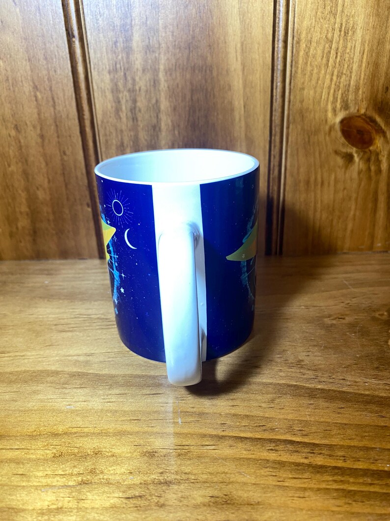 May include: A ceramic mug with a white handle and interior. The exterior features a dark blue background with a celestial design including stars, a crescent moon, and a yellow shape. The mug is on a wooden surface.