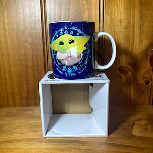 May include: A blue ceramic mug featuring a green character with large eyes and pointed ears. The mug has a white handle and is sitting on a white box. The mug has the text "This is the way" around the character.