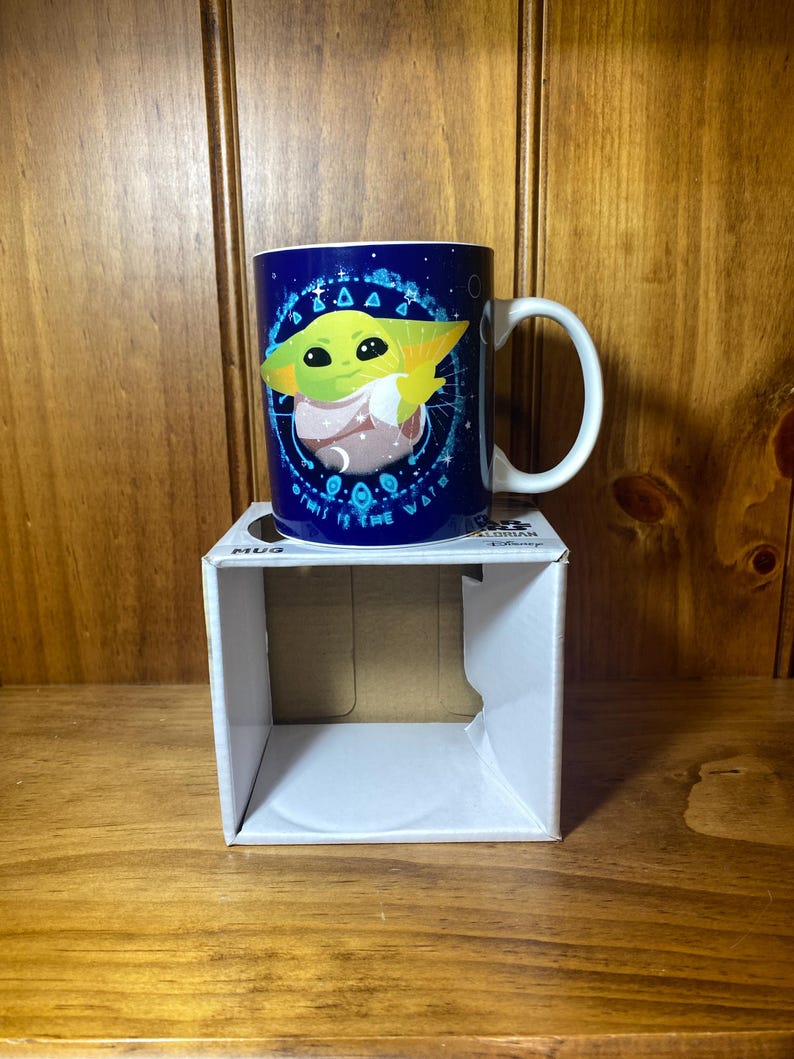 May include: A blue ceramic mug featuring a green character with large eyes and pointed ears. The mug has a white handle and is sitting on a white box. The mug has the text "This is the way" around the character.