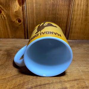 Puede incluir: Una taza de cer&aacute;mica con interior blanco y exterior amarillo que presenta un gr&aacute;fico de silueta marr&oacute;n de un personaje y el texto THE MANDALORIAN impreso en marr&oacute;n. La taza tiene un asa curva en el lateral y una forma cil&iacute;ndrica.