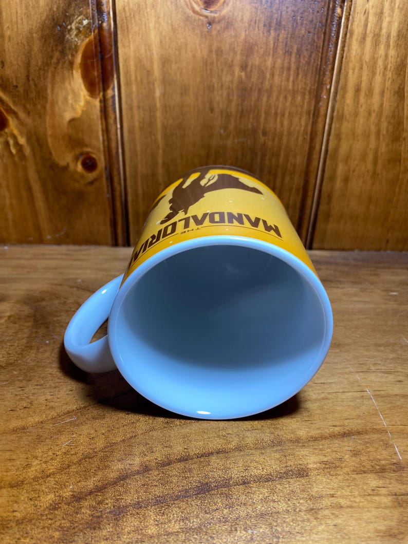 Puede incluir: Una taza de cer&aacute;mica con interior blanco y exterior amarillo que presenta un gr&aacute;fico de silueta marr&oacute;n de un personaje y el texto THE MANDALORIAN impreso en marr&oacute;n. La taza tiene un asa curva en el lateral y una forma cil&iacute;ndrica.