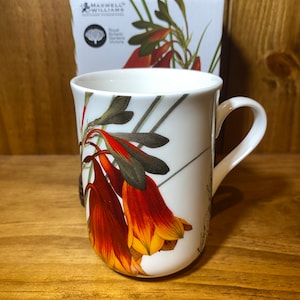 May include: White ceramic mug with a floral design featuring orange and yellow flowers. The mug has a black handle and is sitting on a wooden surface. The text "Maxwell Williams" is visible on a box in the background.