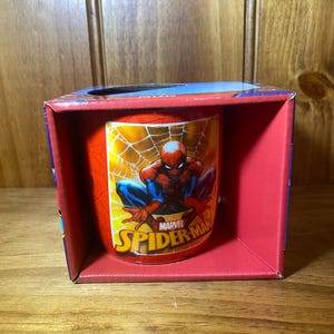 Puede incluir: Una taza roja y amarilla de Spider-Man dentro de una caja roja. La taza presenta a Spider-Man en una telara&ntilde;a con el logotipo de Marvel Spider-Man. La caja tiene una parte superior azul y est&aacute; sobre una superficie de madera.