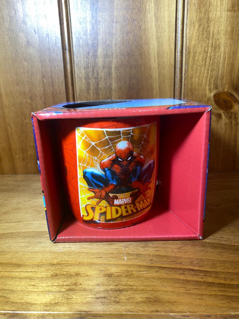 Puede incluir: Una taza roja y amarilla de Spider-Man dentro de una caja roja. La taza presenta a Spider-Man en una telara&ntilde;a con el logotipo de Marvel Spider-Man. La caja tiene una parte superior azul y est&aacute; sobre una superficie de madera.
