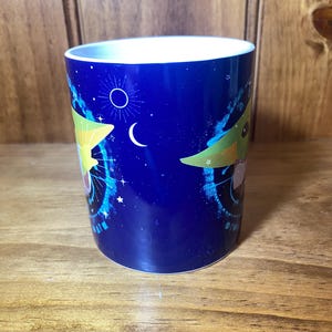 May include: A blue ceramic mug featuring a cartoon character design. The mug has a white interior and a wrap-around design with stars, a crescent moon, and a circular emblem. The design includes a cartoon character with large ears.