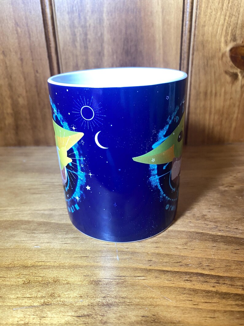 May include: A blue ceramic mug featuring a cartoon character design. The mug has a white interior and a wrap-around design with stars, a crescent moon, and a circular emblem. The design includes a cartoon character with large ears.