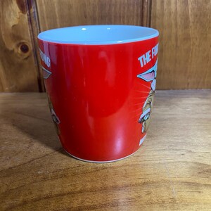 Pode incluir: Uma caneca de cer&acirc;mica vermelha brilhante com um gr&aacute;fico de uma pequena criatura verde de orelhas grandes usando um gorro de Papai Noel. O texto na caneca diz THE FORCE IS STRONG WITH THIS ONE em letras brancas. O interior &eacute; branco.