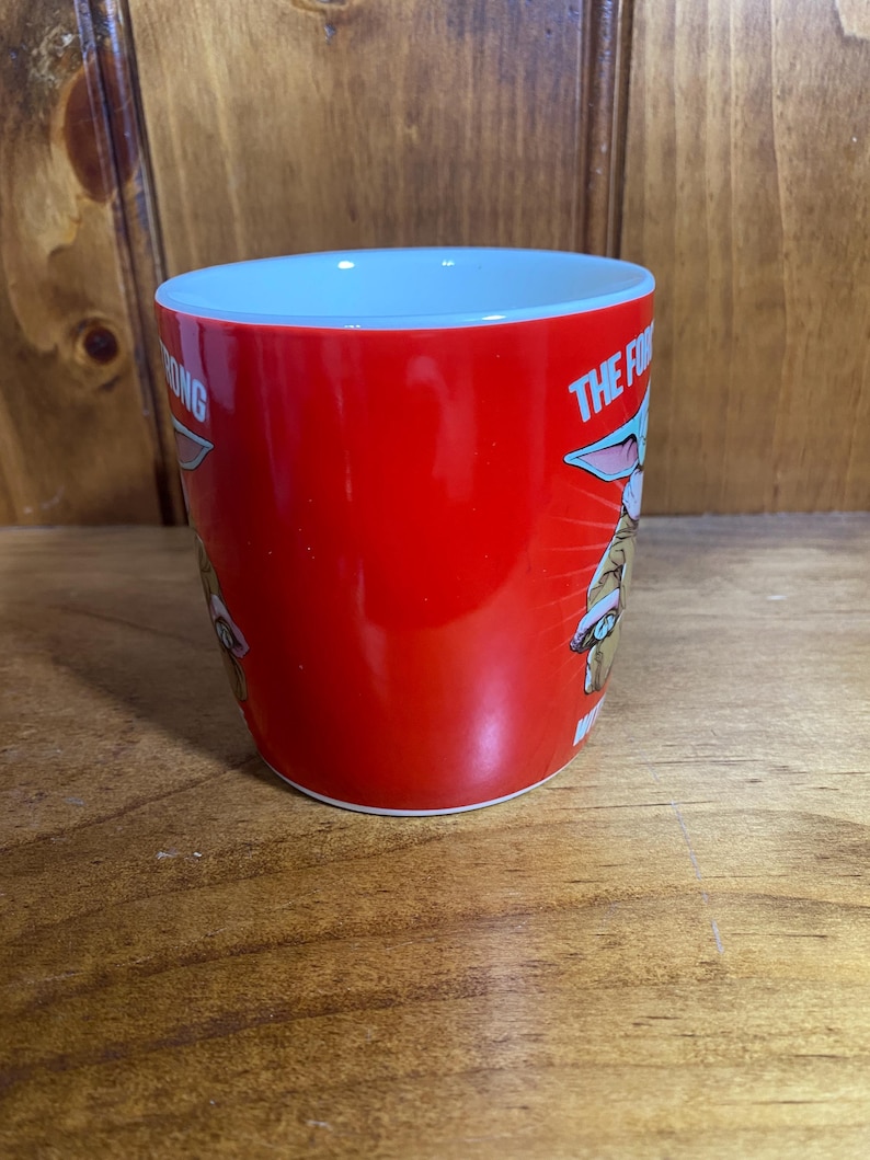 Pode incluir: Uma caneca de cer&acirc;mica vermelha brilhante com um gr&aacute;fico de uma pequena criatura verde de orelhas grandes usando um gorro de Papai Noel. O texto na caneca diz THE FORCE IS STRONG WITH THIS ONE em letras brancas. O interior &eacute; branco.