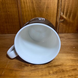 May include: A white ceramic coffee mug with a brown exterior. The mug has a white interior and a curved handle. The mug is sitting on a wooden surface.