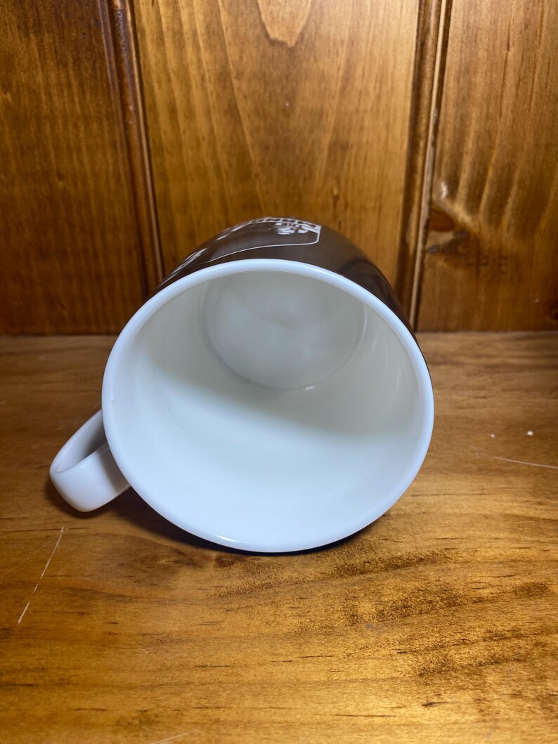 May include: A white ceramic coffee mug with a brown exterior. The mug has a white interior and a curved handle. The mug is sitting on a wooden surface.