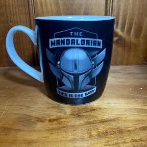 May include: Black ceramic mug with a white handle and rim. The mug features the text "THE MANDALORIAN" above an image of a helmet and the phrase "THIS IS THE WAY".