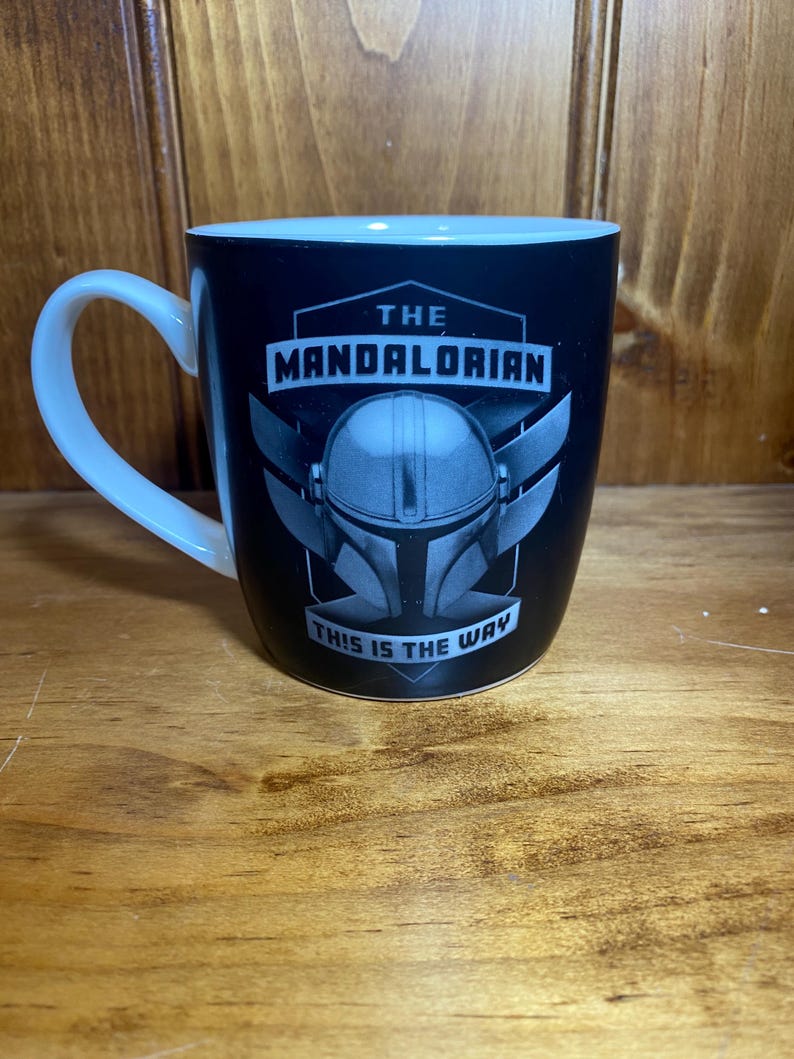 May include: Black ceramic mug with a white handle and rim. The mug features the text "THE MANDALORIAN" above an image of a helmet and the phrase "THIS IS THE WAY".