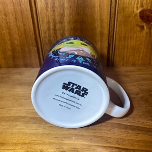 May include: A ceramic Star Wars mug featuring Grogu, also known as Baby Yoda, on a dark blue background. The mug has a white handle and a white base with the Star Wars logo and licensing information. Microwave and dishwasher safe.