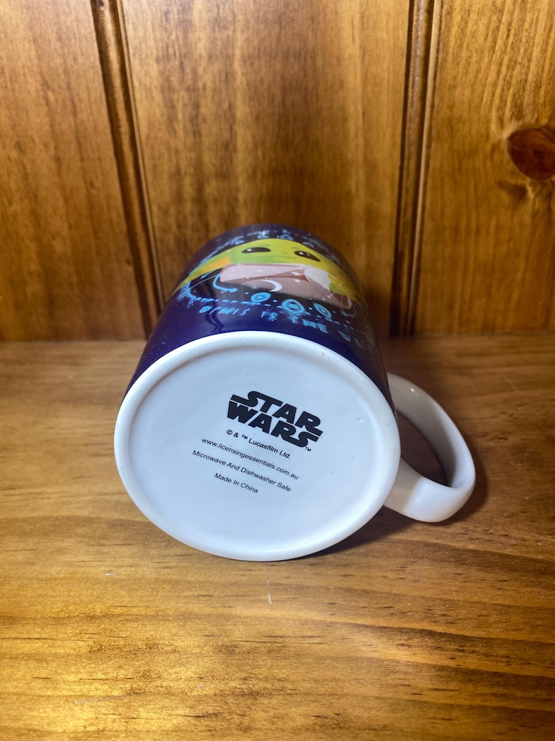 May include: A ceramic Star Wars mug featuring Grogu, also known as Baby Yoda, on a dark blue background. The mug has a white handle and a white base with the Star Wars logo and licensing information. Microwave and dishwasher safe.