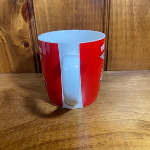Pode incluir: Uma caneca de cer&acirc;mica branca com uma faixa exterior vermelha brilhante. A caneca tem uma pega branca curva e um interior branco. O texto vis&iacute;vel na superf&iacute;cie vermelha inclui a palavra THE em letras mai&uacute;sculas brancas, parcialmente oculta.