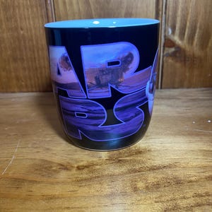 K&ouml;nnte beinhalten: Schwarze Keramik-Tasse mit dem Star Wars-Logo in Lila und Wei&szlig;. Die Tasse hat einen hellblauen Innenraum und eine Landschaftsszene in den Buchstaben. Die Tasse ist zylindrisch und steht auf einer Holzoberfl&auml;che.