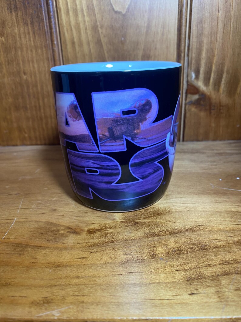 K&ouml;nnte beinhalten: Schwarze Keramik-Tasse mit dem Star Wars-Logo in Lila und Wei&szlig;. Die Tasse hat einen hellblauen Innenraum und eine Landschaftsszene in den Buchstaben. Die Tasse ist zylindrisch und steht auf einer Holzoberfl&auml;che.