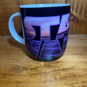 K&ouml;nnte beinhalten: Eine Keramiktasse mit schwarzer Au&szlig;enseite und dem Wort "STAR" in gro&szlig;en, violetten, stilisierten Buchstaben. Die Buchstaben sind auf einer Sonnenuntergangsszene mit Lila-, Rosa- und Oranget&ouml;nen &uuml;berlagert. Der Griff und das Innere sind hellblau.