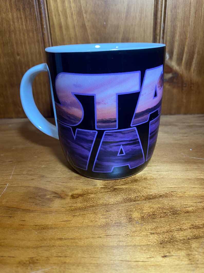 K&ouml;nnte beinhalten: Eine Keramiktasse mit schwarzer Au&szlig;enseite und dem Wort "STAR" in gro&szlig;en, violetten, stilisierten Buchstaben. Die Buchstaben sind auf einer Sonnenuntergangsszene mit Lila-, Rosa- und Oranget&ouml;nen &uuml;berlagert. Der Griff und das Innere sind hellblau.