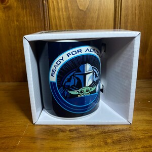 K&ouml;nnte beinhalten: Dunkelblaue Keramiktasse mit einer Grafik von The Mandalorian und Grogu, mit dem Text "READY FOR ADVENTURE" in einem kreisf&ouml;rmigen Design. Die Tasse wird in einer wei&szlig;en Box pr&auml;sentiert.