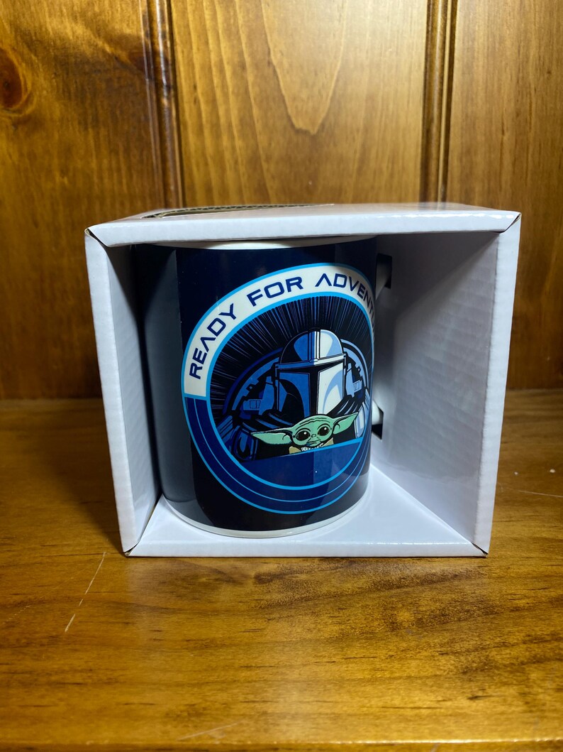 K&ouml;nnte beinhalten: Dunkelblaue Keramiktasse mit einer Grafik von The Mandalorian und Grogu, mit dem Text "READY FOR ADVENTURE" in einem kreisf&ouml;rmigen Design. Die Tasse wird in einer wei&szlig;en Box pr&auml;sentiert.