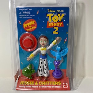 Puede incluir: Un paquete de plástico que contiene una muñeca de Jessie de la película Toy Story 2. La muñeca es articulada y viene con un sombrero, una serpiente, una tortuga y un pájaro. El paquete es azul y amarillo con las palabras "Toy Story 2" y "Jessie & Critters" impresas en él.