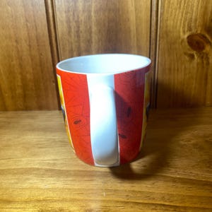 Puede incluir: Una taza de caf&eacute; de cer&aacute;mica con un asa blanca y un dise&ntilde;o a rayas rojas y blancas. La taza tiene un interior blanco y un dise&ntilde;o de telara&ntilde;a. La taza est&aacute; sobre una superficie de madera.
