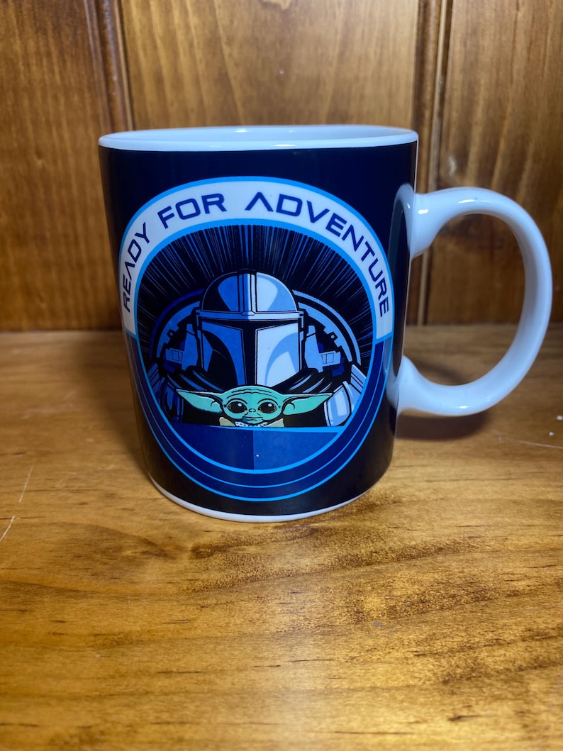 K&ouml;nnte beinhalten: Eine wei&szlig;e Keramiktasse mit blauem &Auml;u&szlig;eren, die den Text "READY FOR ADVENTURE" um ein Bild eines Mandalorianer-Helms und Grogu tr&auml;gt. Die Tasse hat einen wei&szlig;en Henkel und ein wei&szlig;es Inneres.