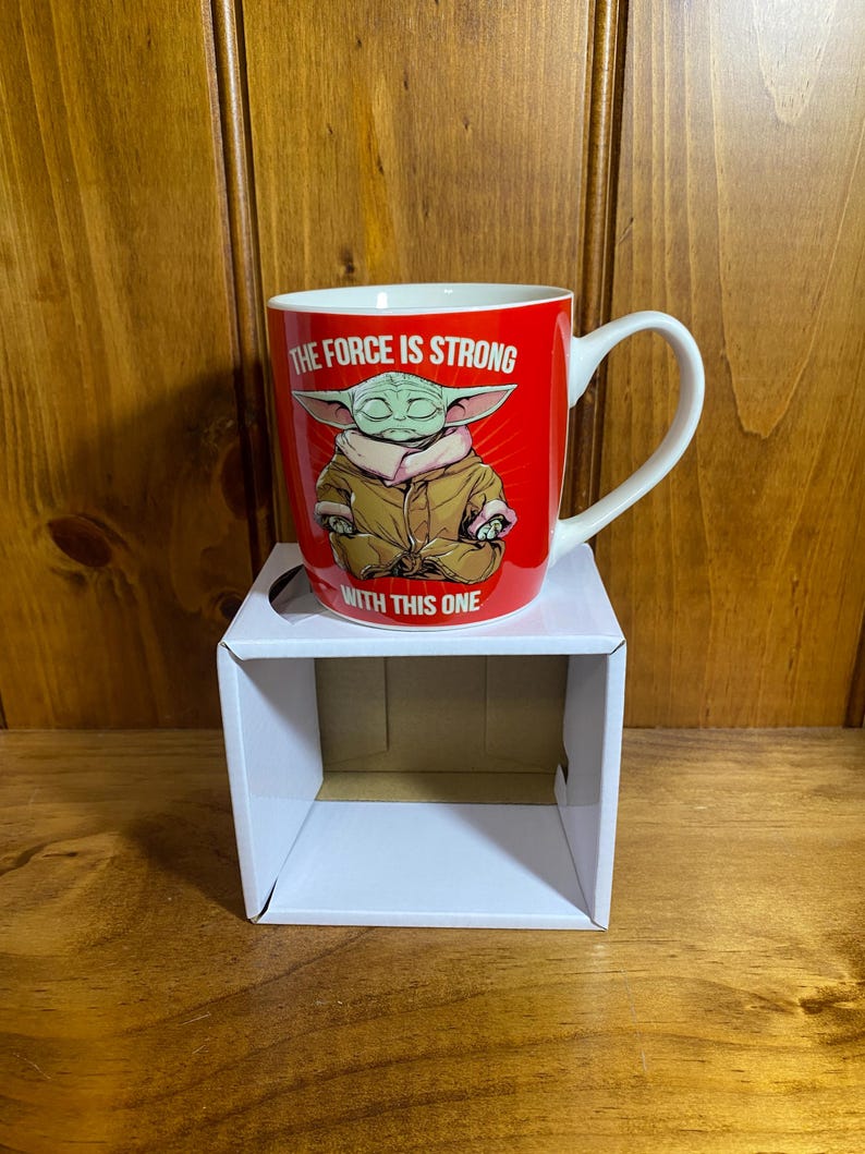 Pode incluir: Uma caneca de cer&acirc;mica vermelha com interior e al&ccedil;a brancos, apresentando um gr&aacute;fico de uma pequena criatura verde com uma t&uacute;nica bege. O texto "THE FORCE IS STRONG" aparece acima do personagem e "WITH THIS ONE" est&aacute; impresso abaixo.