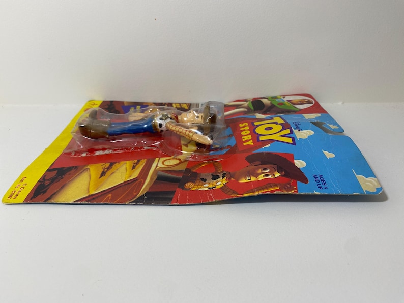 以下が含まれることがあります： トイストーリーのウッディのプラスチック製アクションフィギュア。クラシックなカウボーイの衣装を着て、投げ縄を持っています。フィギュアは、トイストーリーのロゴと「Ages 4 and Up」の文字が書かれたカラフルなカードにパッケージされています。