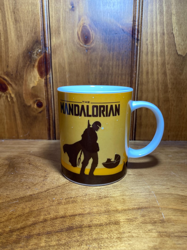 Puede incluir: Una taza de cer&aacute;mica blanca con fondo amarillo y una silueta negra de The Mandalorian y Grogu en una c&aacute;psula flotante. El texto THE MANDALORIAN est&aacute; impreso en negro en la parte superior. La taza tiene un asa blanca y una forma cil&iacute;ndrica.