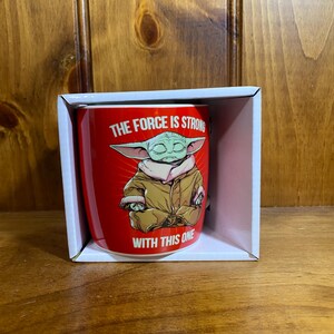 Pode incluir: Uma caneca de cer&acirc;mica vermelha com uma ilustra&ccedil;&atilde;o gr&aacute;fica de uma criatura verde de orelhas grandes vestindo um manto bege, sentada em posi&ccedil;&atilde;o de medita&ccedil;&atilde;o. O texto "THE FORCE IS STRONG WITH THIS ONE" est&aacute; impresso em letras brancas.
