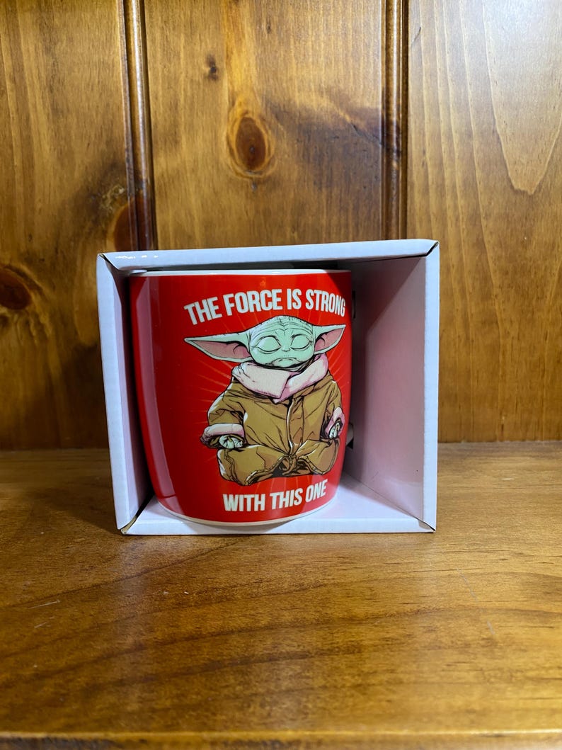 Pode incluir: Uma caneca de cer&acirc;mica vermelha com uma ilustra&ccedil;&atilde;o gr&aacute;fica de uma criatura verde de orelhas grandes vestindo um manto bege, sentada em posi&ccedil;&atilde;o de medita&ccedil;&atilde;o. O texto "THE FORCE IS STRONG WITH THIS ONE" est&aacute; impresso em letras brancas.