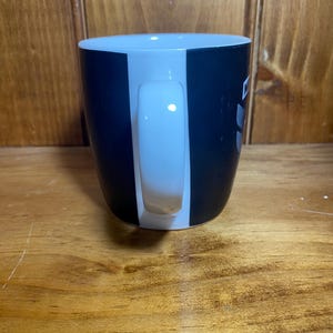 May include: A black and white ceramic coffee mug with a white handle. The mug has a vertical white stripe on the front and a black stripe on the back. The mug is sitting on a wooden surface.