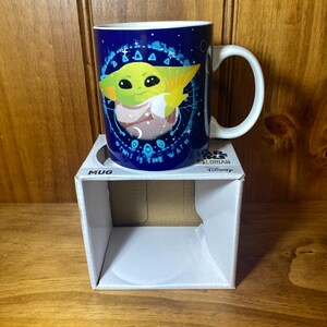 May include: A blue ceramic mug featuring a green character with large eyes and pointed ears. The mug has a white handle and a design that includes the text "THIS IS THE WAY". The mug is on a white box labeled "MUG".