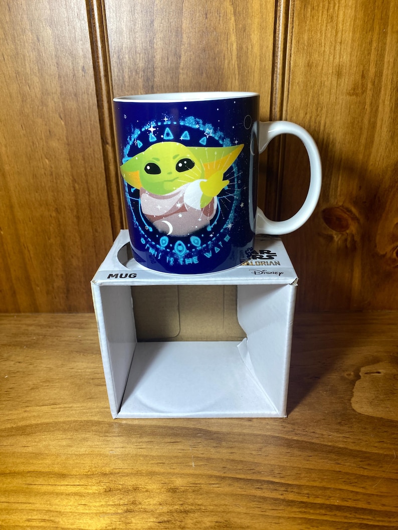 May include: A blue ceramic mug featuring a green character with large eyes and pointed ears. The mug has a white handle and a design that includes the text "THIS IS THE WAY". The mug is on a white box labeled "MUG".