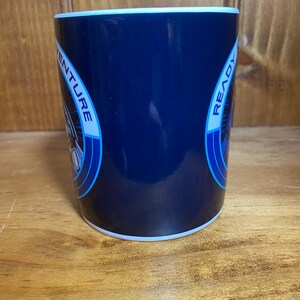K&ouml;nnte beinhalten: Eine marineblaue Keramiktasse mit wei&szlig;em Rand und Boden. Die Tasse tr&auml;gt die Worte "ADVENTURE READY" in einem kreisf&ouml;rmigen Design mit blauen Akzenten. Die Tasse ist zylindrisch und steht auf einer Holzoberfl&auml;che.