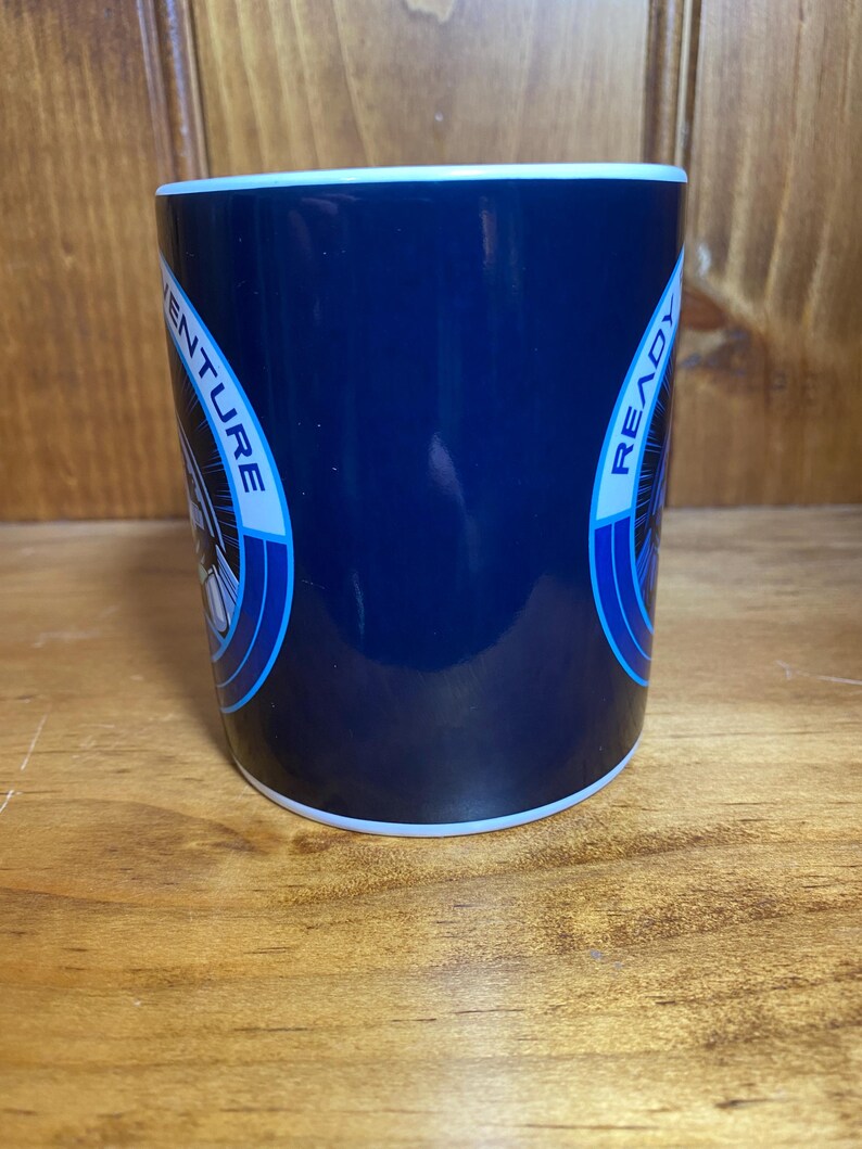 K&ouml;nnte beinhalten: Eine marineblaue Keramiktasse mit wei&szlig;em Rand und Boden. Die Tasse tr&auml;gt die Worte "ADVENTURE READY" in einem kreisf&ouml;rmigen Design mit blauen Akzenten. Die Tasse ist zylindrisch und steht auf einer Holzoberfl&auml;che.