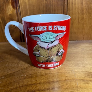 Pode incluir: Uma caneca de cer&acirc;mica vermelha brilhante com um gr&aacute;fico de um personagem verde de orelhas grandes em um manto bege. O texto "THE FORCE IS STRONG" est&aacute; impresso em branco acima e "WITH THIS ONE" est&aacute; impresso em branco abaixo.