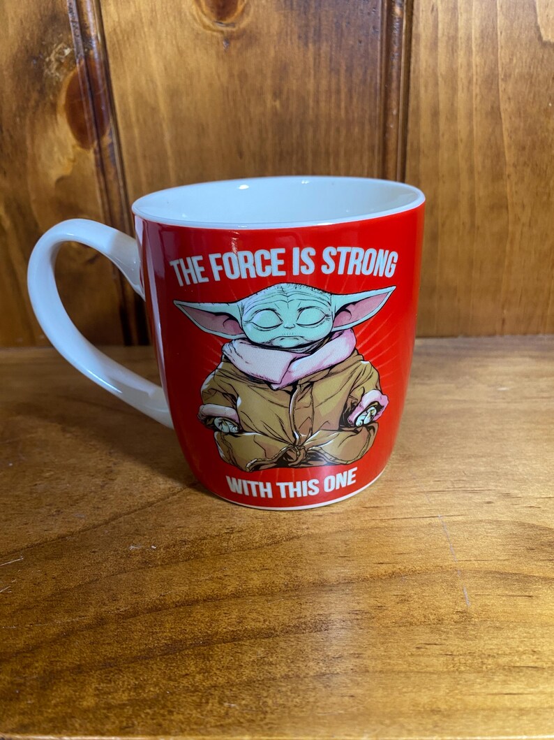 Pode incluir: Uma caneca de cer&acirc;mica vermelha brilhante com um gr&aacute;fico de um personagem verde de orelhas grandes em um manto bege. O texto "THE FORCE IS STRONG" est&aacute; impresso em branco acima e "WITH THIS ONE" est&aacute; impresso em branco abaixo.