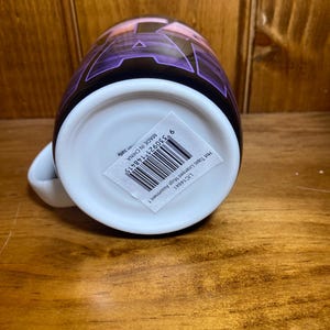 K&ouml;nnte beinhalten: Eine wei&szlig;e Keramiktasse mit einem lila und schwarzen Design. Die Tasse hat einen wei&szlig;en Henkel und einen Aufkleber mit Barcode und Text. Die Tasse steht auf einer Holzoberfl&auml;che.