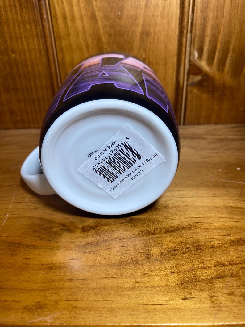 K&ouml;nnte beinhalten: Eine wei&szlig;e Keramiktasse mit einem lila und schwarzen Design. Die Tasse hat einen wei&szlig;en Henkel und einen Aufkleber mit Barcode und Text. Die Tasse steht auf einer Holzoberfl&auml;che.