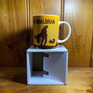 Puede incluir: Una taza de cer&aacute;mica blanca con un dise&ntilde;o gr&aacute;fico amarillo y el texto THE MANDALORIAN. La ilustraci&oacute;n muestra la silueta oscura de un personaje con armadura de pie junto a una peque&ntilde;a c&aacute;psula flotante. La taza tiene asa e interior blancos.