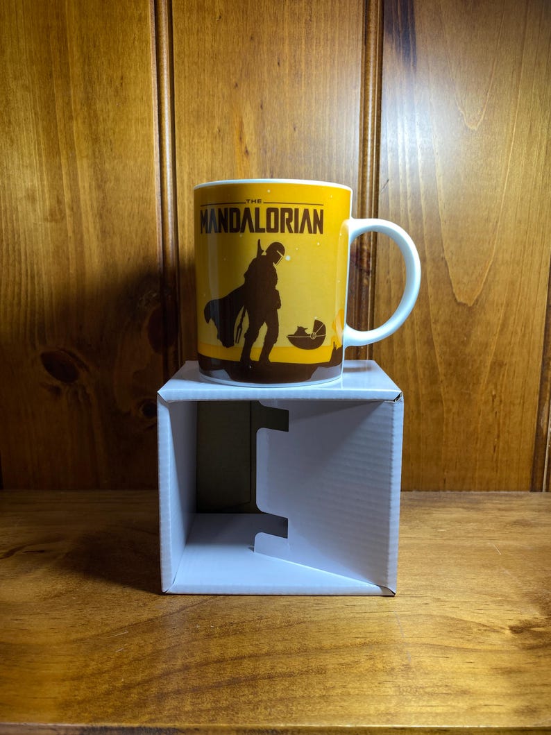 Puede incluir: Una taza de cer&aacute;mica blanca con un dise&ntilde;o gr&aacute;fico amarillo y el texto THE MANDALORIAN. La ilustraci&oacute;n muestra la silueta oscura de un personaje con armadura de pie junto a una peque&ntilde;a c&aacute;psula flotante. La taza tiene asa e interior blancos.