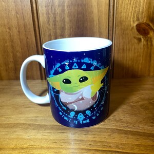 May include: A white-handled ceramic mug with a dark blue background featuring a green alien character. The character is surrounded by a circular design with the text "THIS IS THE WAY". The mug has a white interior.