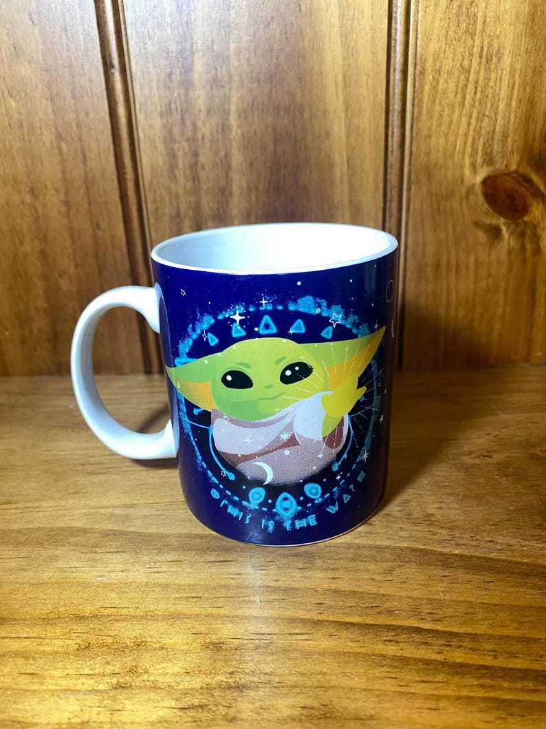 May include: A white-handled ceramic mug with a dark blue background featuring a green alien character. The character is surrounded by a circular design with the text "THIS IS THE WAY". The mug has a white interior.