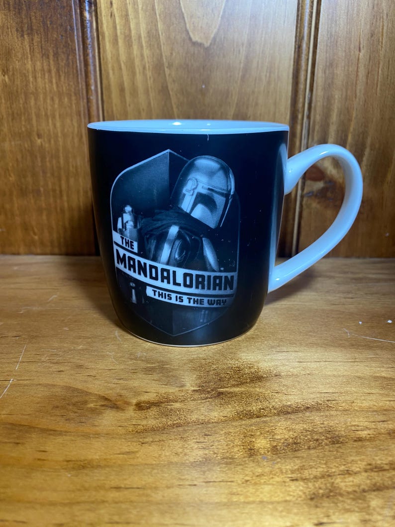 May include: Black ceramic mug with a white handle and interior. The mug features a black and white image of The Mandalorian with the text "THE MANDALORIAN THIS IS THE WAY" in white.