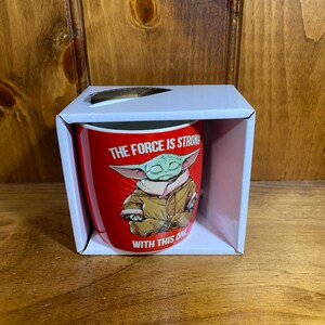 Pode incluir: Uma caneca de cer&acirc;mica vermelha dentro de uma caixa de papel&atilde;o branca. A caneca apresenta um gr&aacute;fico de um personagem verde de orelhas grandes com um manto bege, com o texto branco THE FORCE IS STRONG WITH THIS ONE impresso ao redor.