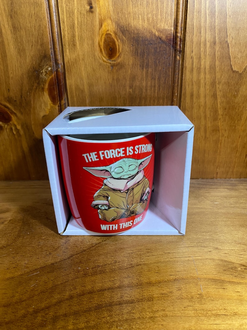 Pode incluir: Uma caneca de cer&acirc;mica vermelha dentro de uma caixa de papel&atilde;o branca. A caneca apresenta um gr&aacute;fico de um personagem verde de orelhas grandes com um manto bege, com o texto branco THE FORCE IS STRONG WITH THIS ONE impresso ao redor.