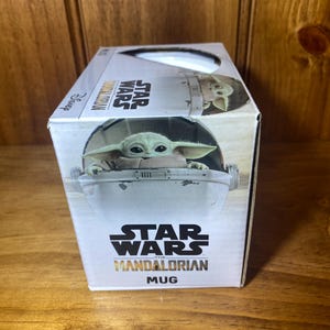 May include: A white box featuring a Star Wars Mandalorian mug. The box has an image of Grogu in his floating pram. The Star Wars logo and the word "Mug" are printed on the box.